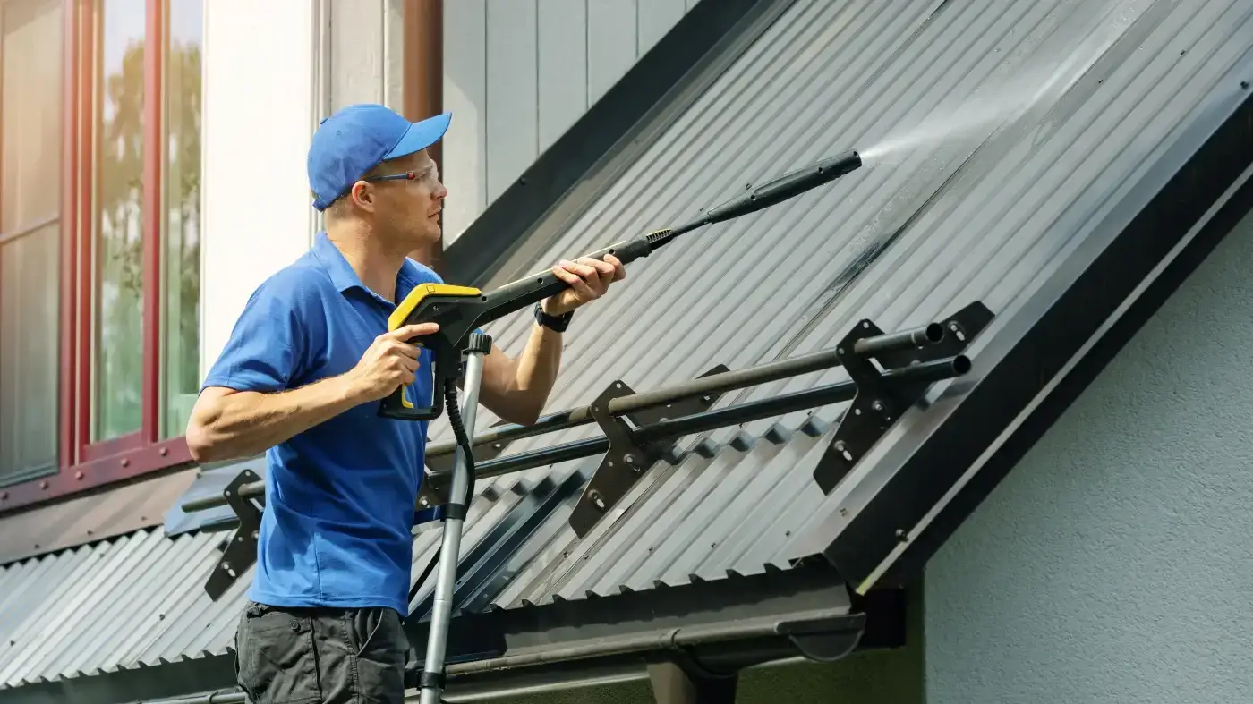 A close view of a roofer on a ladder pressure-washing a building’s metal roof.
