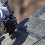 A close view of a roofer nailing an asphalt shingle roof with a nail gun.