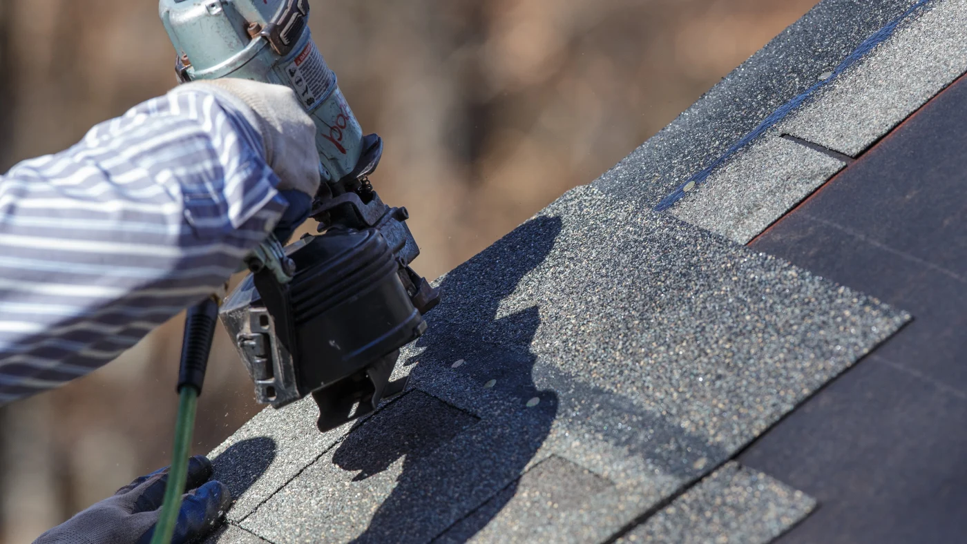 A close view of a roofer nailing an asphalt shingle roof with a nail gun.