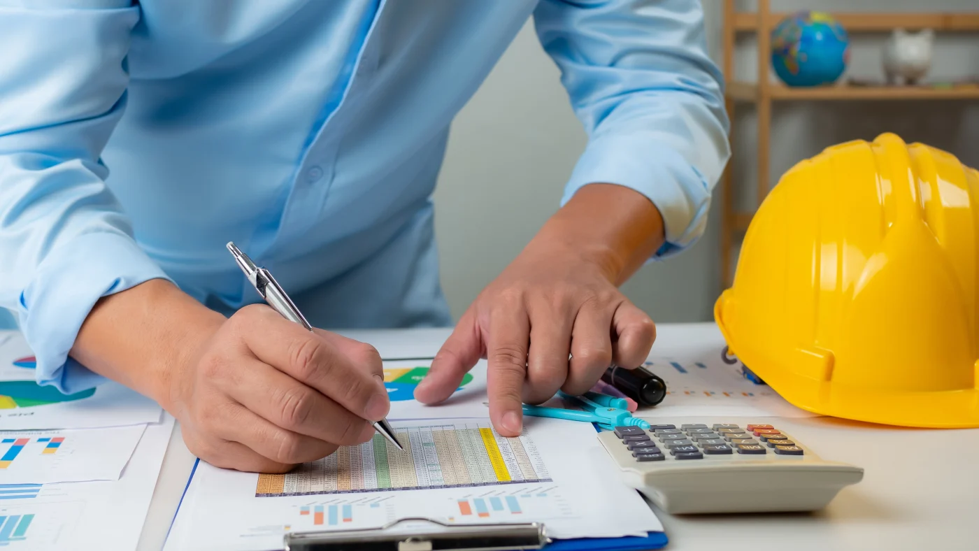 A close view of a man a table looking over a sheet of numbers with a calculator and hard hat beside him.