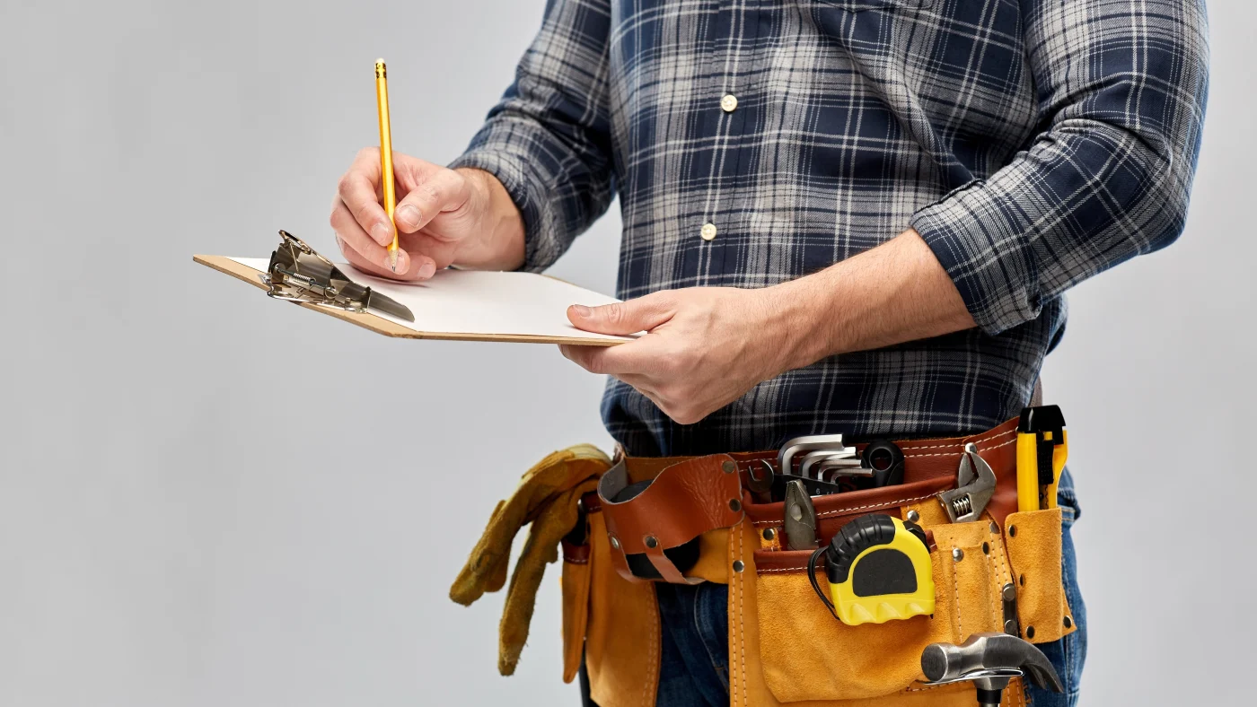 A close view of a man in a flannel shirt with a tool belt around his waist writing on a clipboard he holds in his hand.