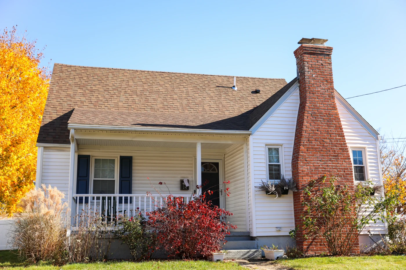A single-family home with a brown shingled roof and a red brick chimney. The house’s siding is white, as is the porch railing, but the window shutters are black, and so is the front door.