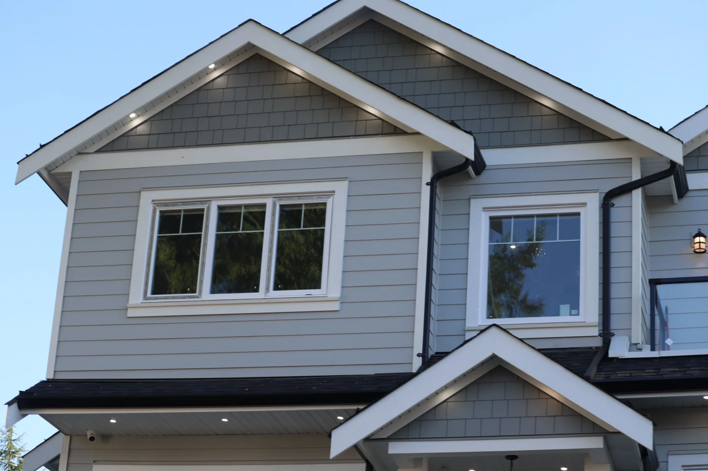 A view from the ground of the upper level of a multi-story home with light gray siding. The gutter system is black, and the home's trim is white.