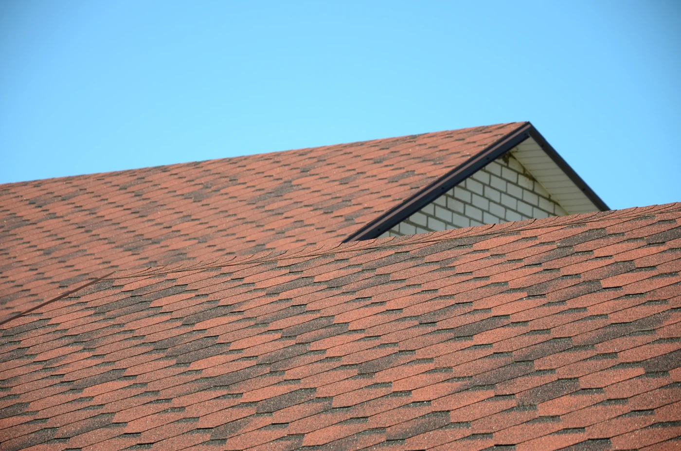 The upper portion of a home’s roof is covered in reddish-brown and black shingles. The sky above the home is bright blue.