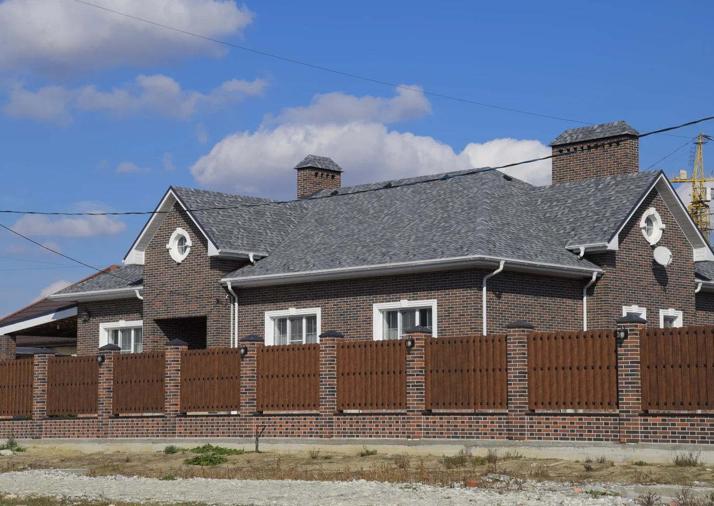 A large brick home surrounded by a low brick wall topped with a dark wooden fence. The home has a dark gray shingled roof, white gutters, and trim.