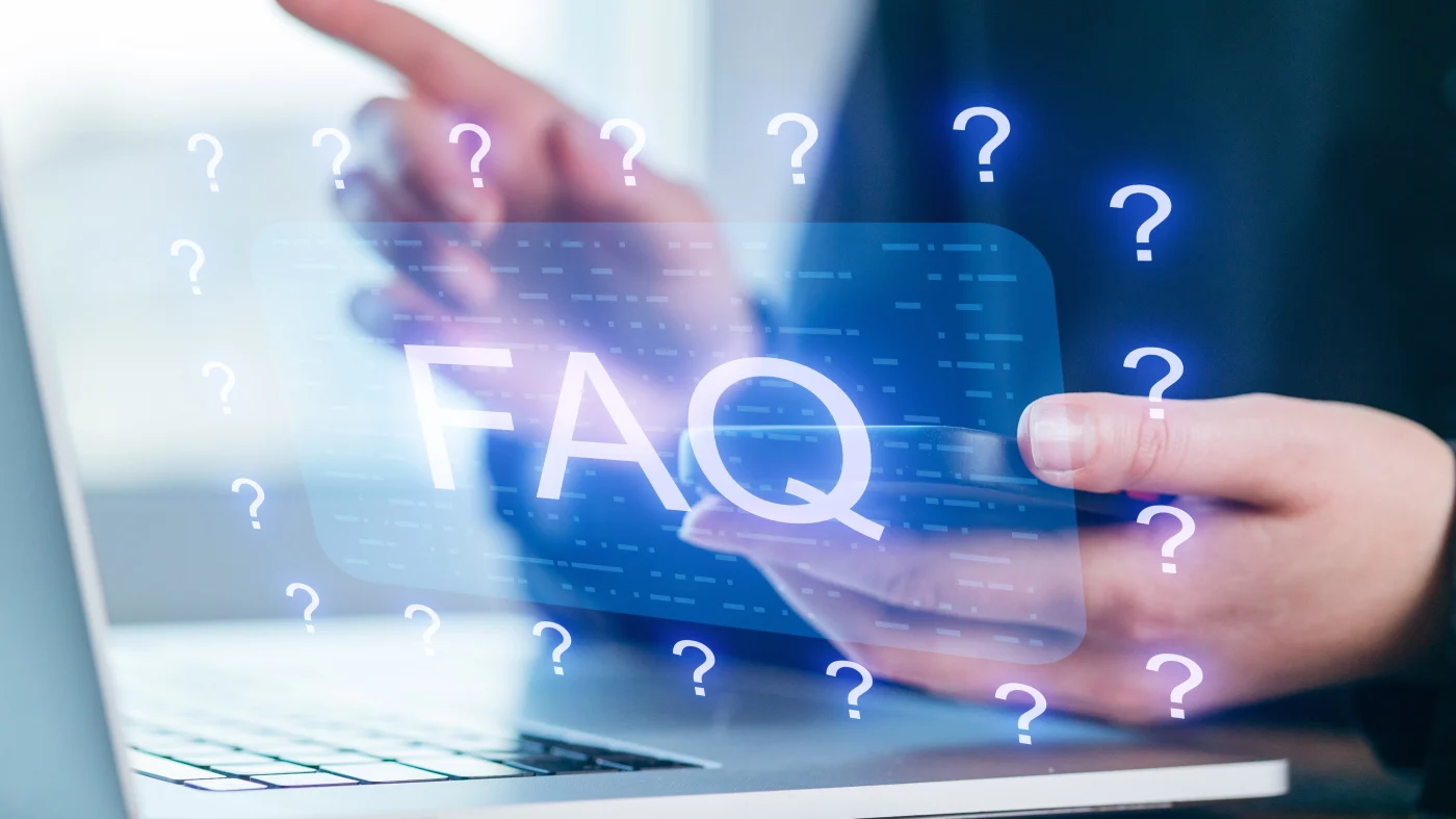 A close view of a woman sitting at a laptop computer with her phone in one hand. Hovering over the image is a graphic that shows the letters FAQ surrounded by question marks.