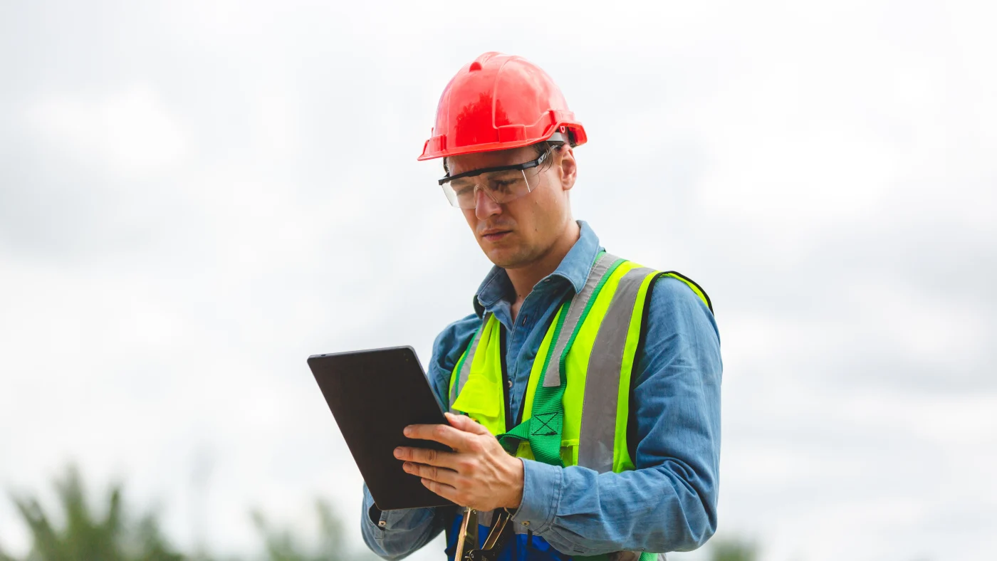 A close view of a roof inspector in full safety gear, marking a checklist on a clipboard he’s holding in his hand.