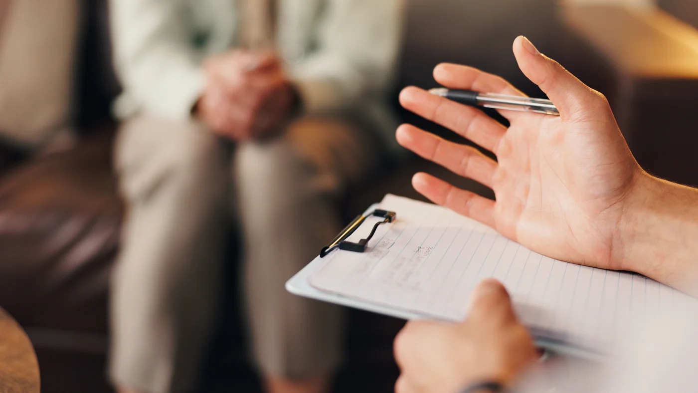 A close-up view of a man with a clipboard talking with a woman sitting on the couch in the background.