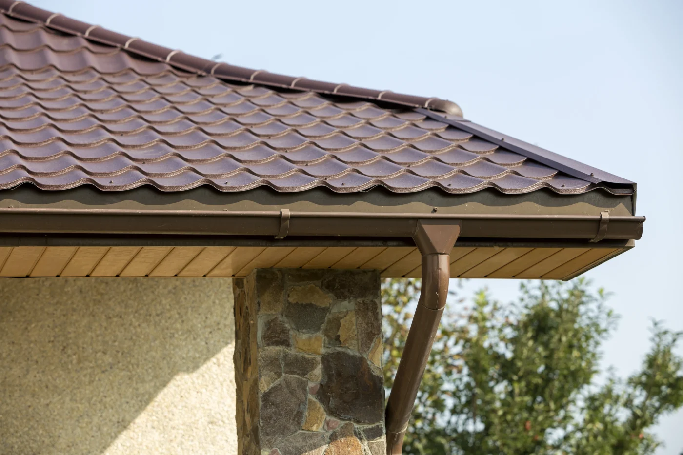 The corner of a home’s roof, with brown shingles and a brown gutter system visible. The house’s exterior is a combination of tan stucco and natural stone.