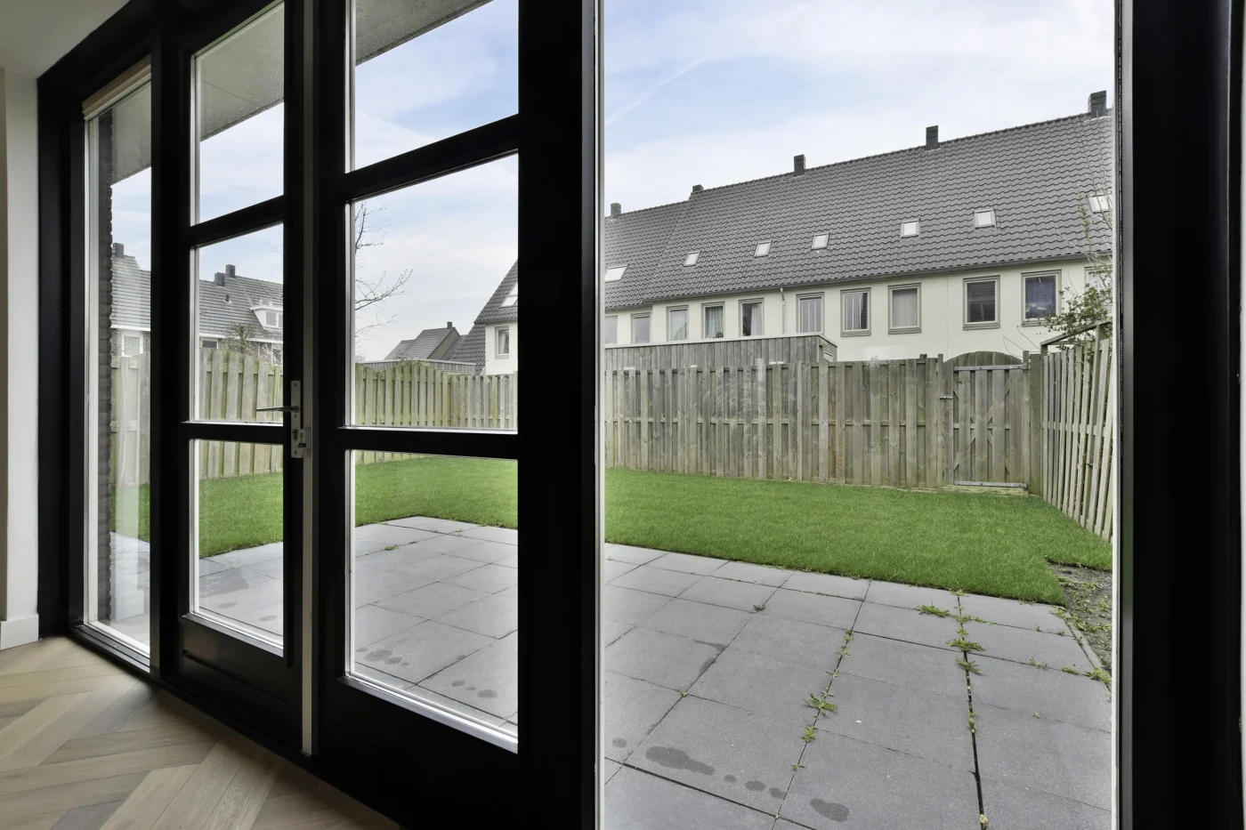A view from inside a home, looking out and into the backyard. There’s a stone-paved patio and a small grassy space. The entire back wall is glass, with a set of French doors in the middle and floor-to-ceiling glass windows on both sides. The window and door frames are black, and the flooring inside the home is wood planking in assorted colors.