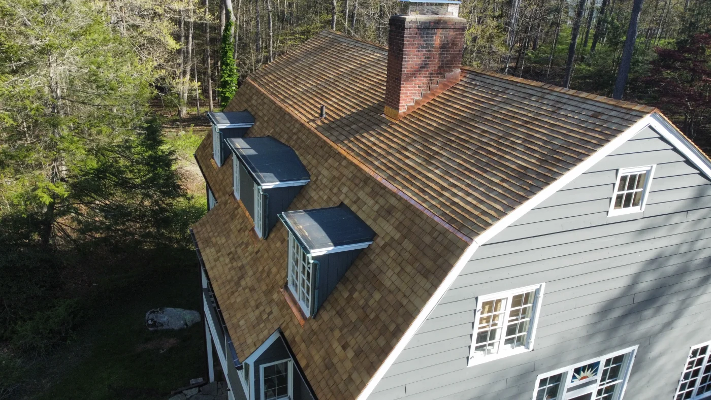 A shot taken with a drone of the side of a home with light gray siding and brown wooden shingles. There are many tall, green trees surrounding the house. The gutters and window frames are white.