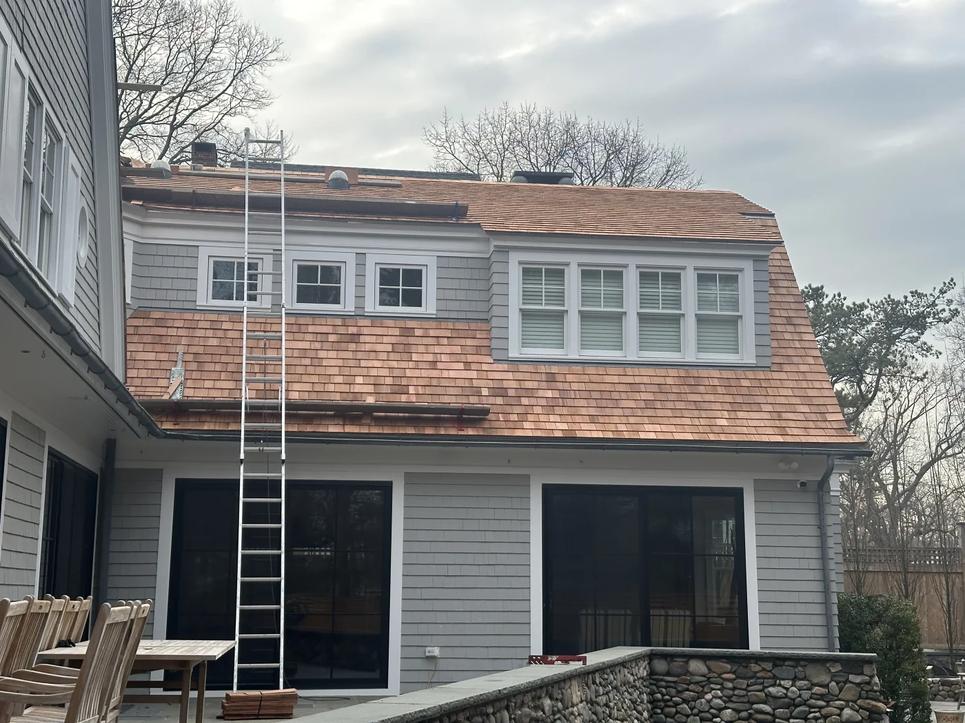 A home that has been given a roof replacement, with the ladder that reaches the upper level still in place. The home’s exterior is covered in light gray siding, and the roof is covered in brown wooden shingles in assorted shades. The gutters are gray, while the window frames and house trim are white.