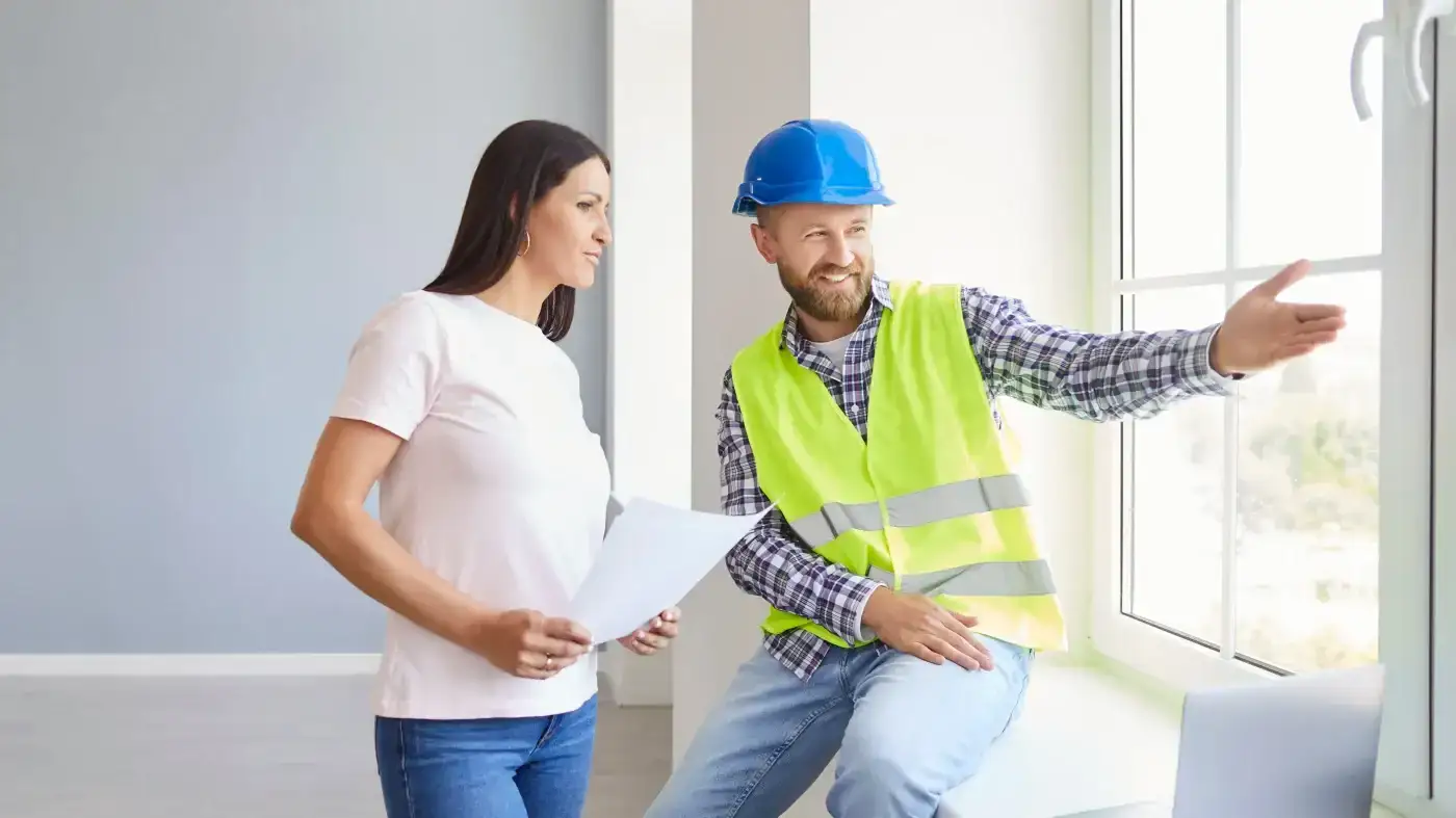 Personalized Window Project Assessment | Fairfield County, CT | Sound Renovation A man in a hard hat and safety vest sits at a window and points toward it while talking with a woman standing next to him.