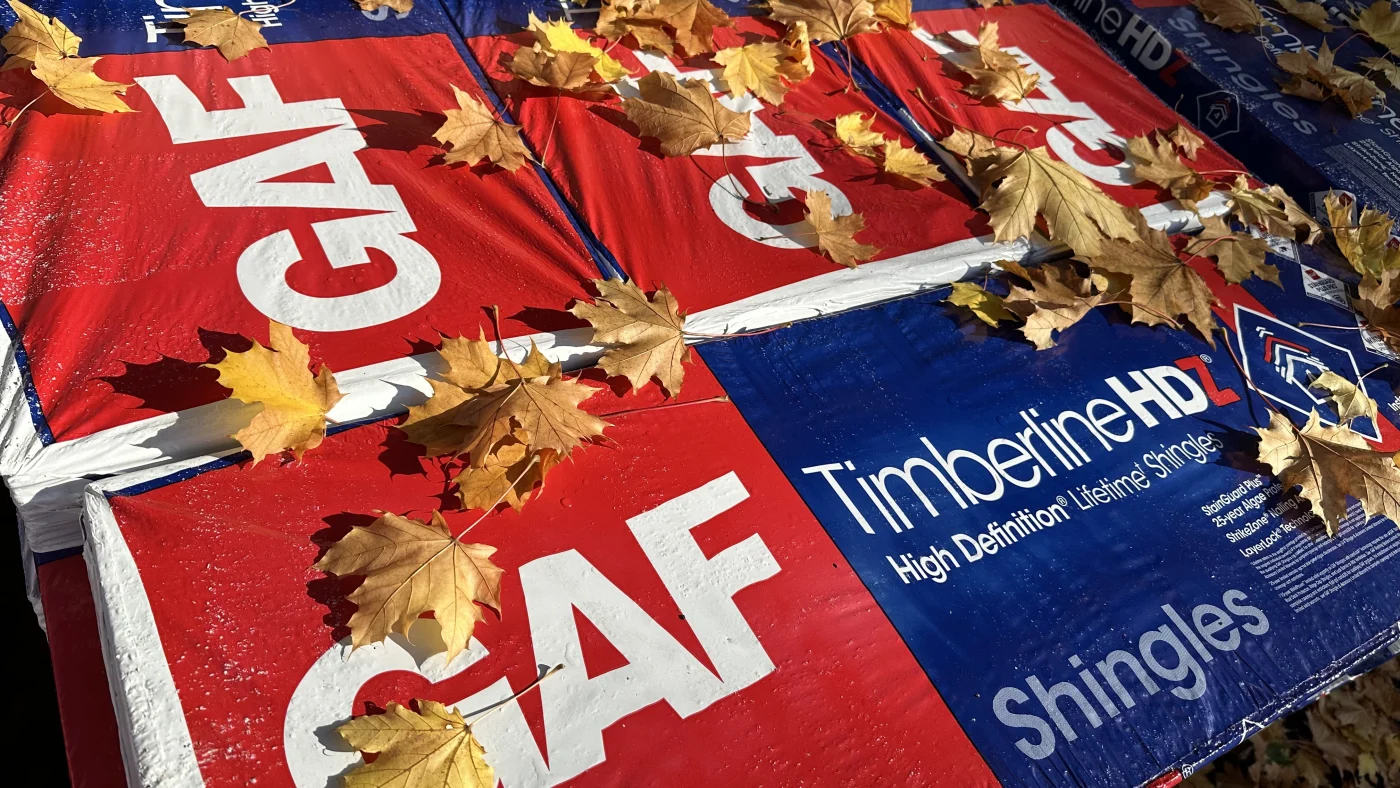 A close-up view of two stacks of GAF Timberline HDZ shingles on a work site with fallen leaves scattered on top of them.