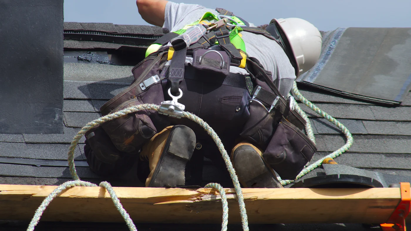 A close view of a roofer in safety gear repairing damage in an asphalt shingle roof.