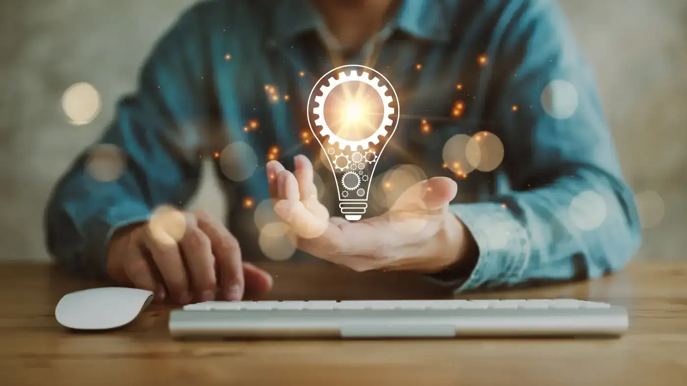 A frontal close-up of a man sitting at a table with a computer keyboard in front of him. His left hand is extended and a vivid lightbulb graphic hovers over it.