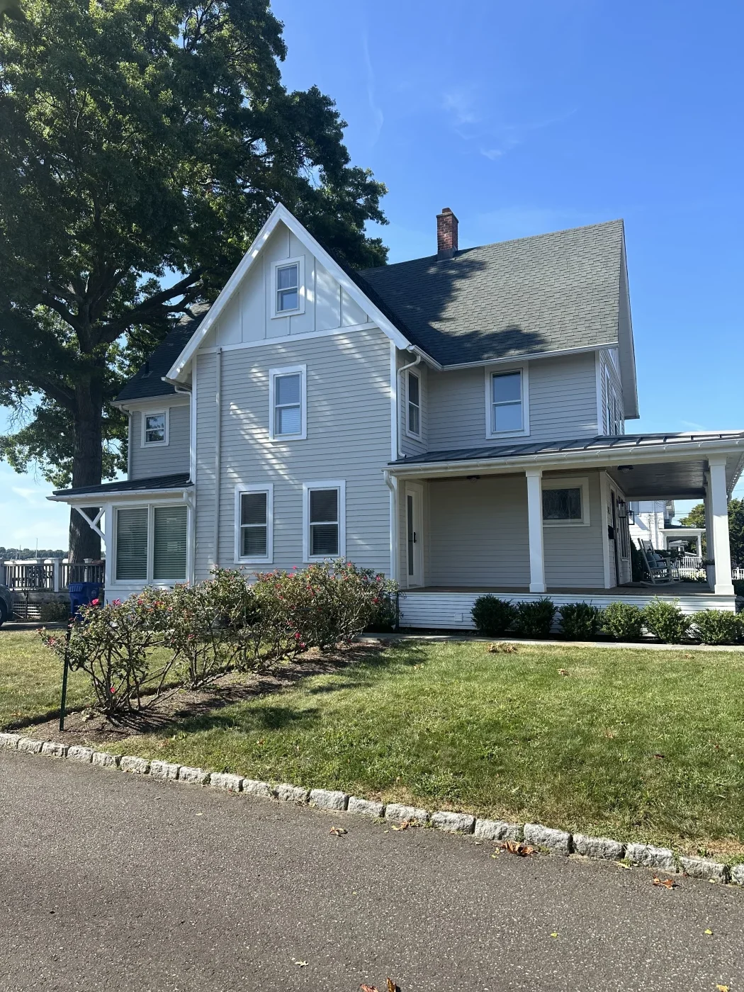Top-Notch Residential Roofing In Wilton, CT | Sound Renovation A multi-story home with a light gray shingle roof on the upper level and gray metal roofing over the lower level and covering the wraparound porch. The home has ta siding and white window frames, gutters, and trim.