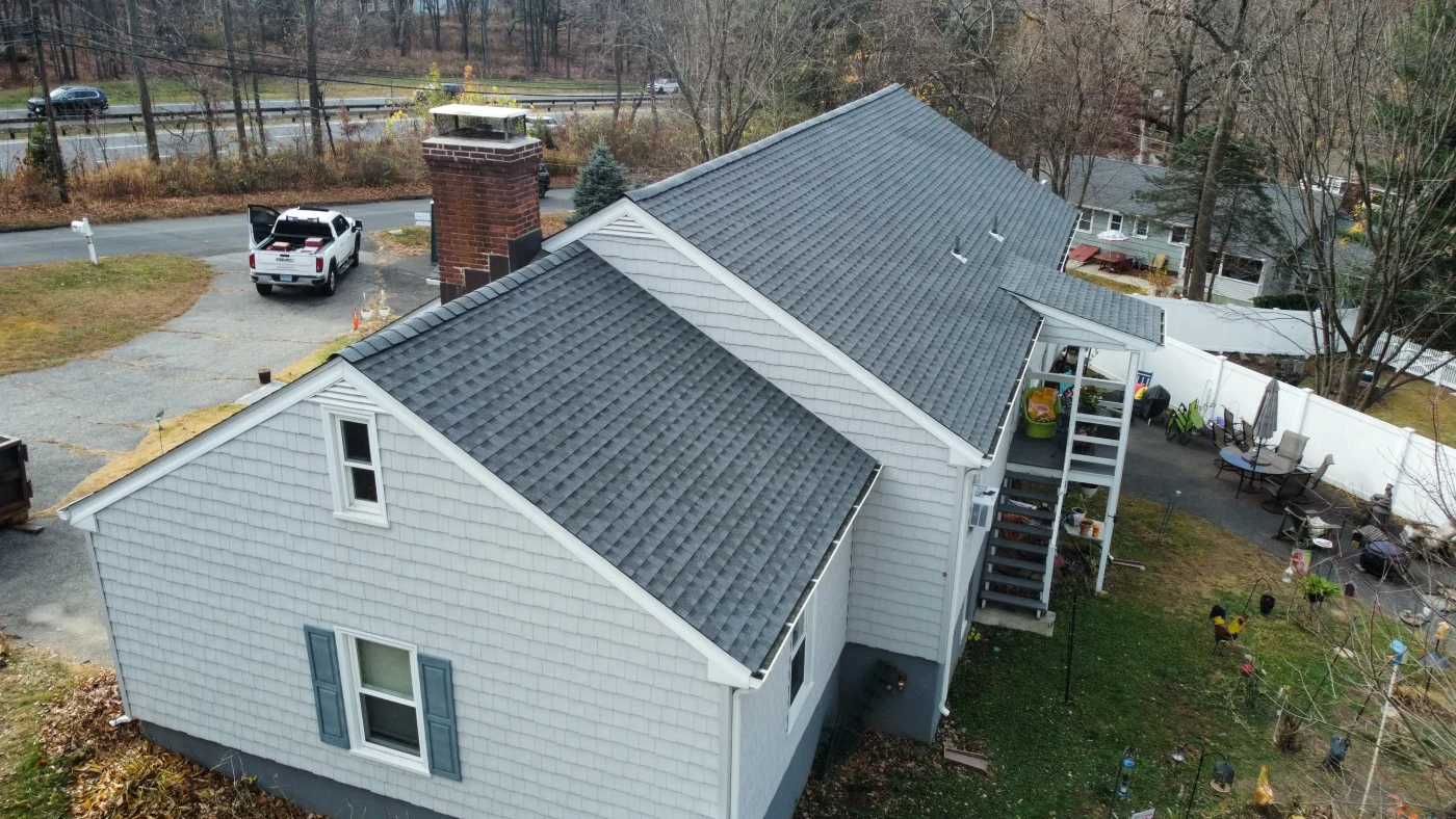 Tranquil Residential Roofing In Wilton, CT | Sound Renovation A drone photo of a home with light blue-gray shingles on the exterior and a gray shingled roof. The gutters are white, and so is the window trim, but the window shutters are blue.
