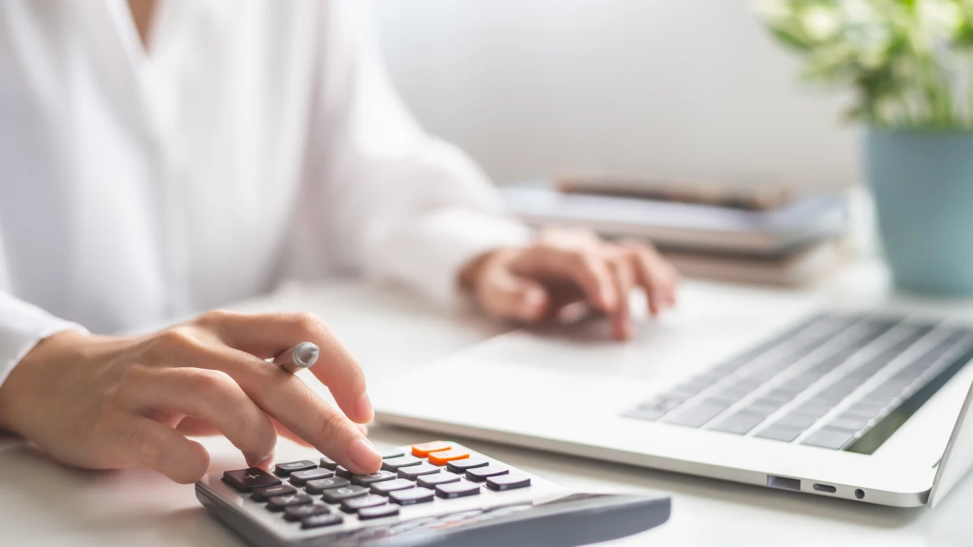 A close view of a woman at a desk simultaneously using a calculator and a laptop computer.