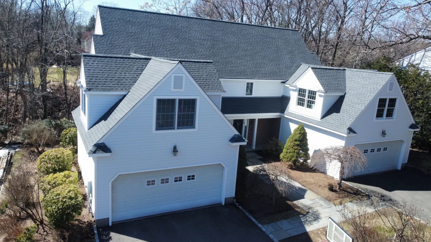 Triumphant Residential Roofing In Wilton, CT | Sound Renovation A photo taken with a drone showing a multi-level home with white siding, a dark gray shingled roof, and a wide, white garage door. There are tall, leafless trees next to and behind the home.