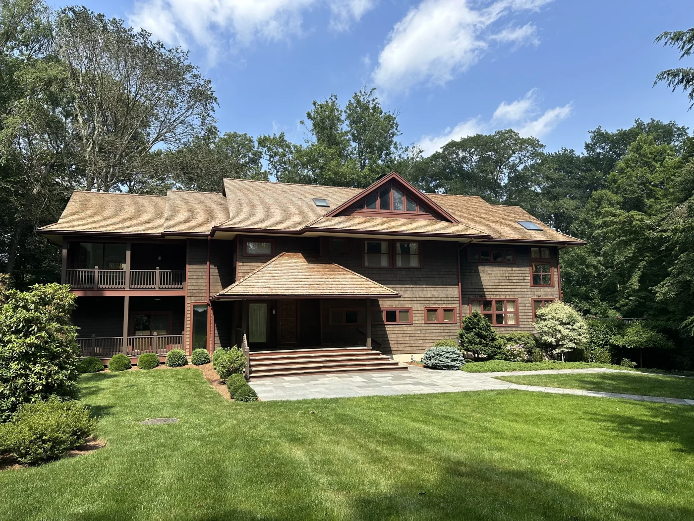 A photo taken from the yard showing a multi-story home with a brown, shingled exterior and a cedar shingle roof. The home has a large, grassy front yard and many trees surrounding it.