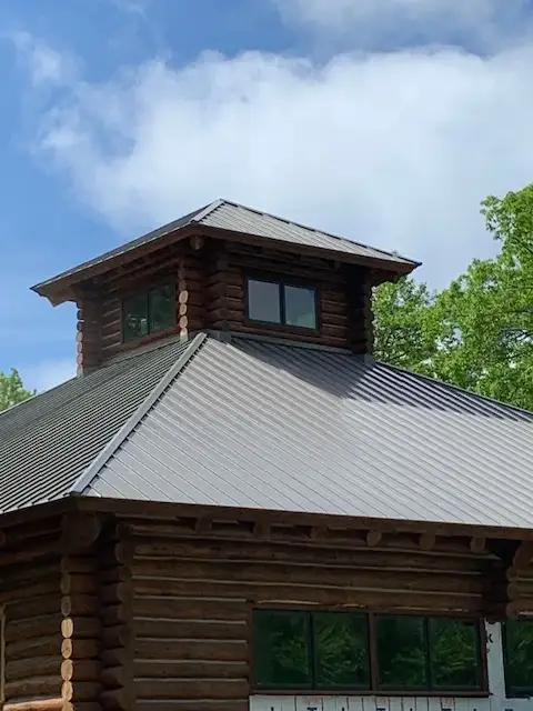 A photo showing a small portion of a home with a wooden log exterior and a dark-colored metal roof. There is a small room on the visible second level of the home, with windows on both sides. The lower level also has multiple windows. Behind the home, the tops of leafy green trees can be seen.
