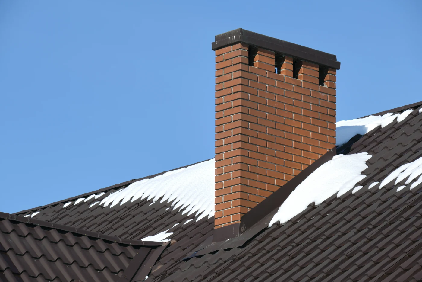 A black metal roof’s peak, with a tall, thin red brick chimney at the center of the image. Above the roof, the sky is bright blue.