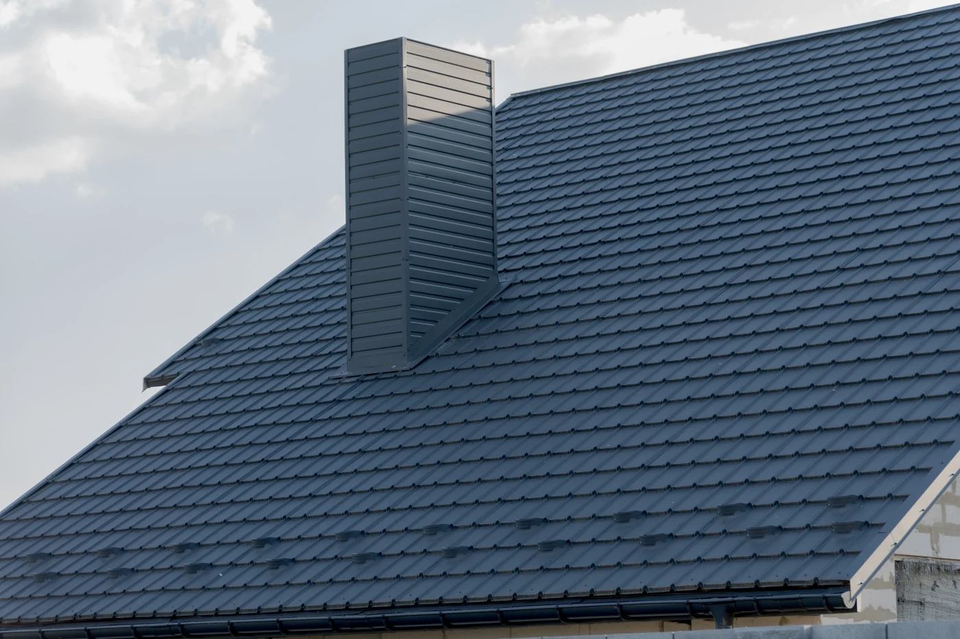 A close-up view of a steep roof covered in dark gray metal. The chimney is also covered in dark gray panels.