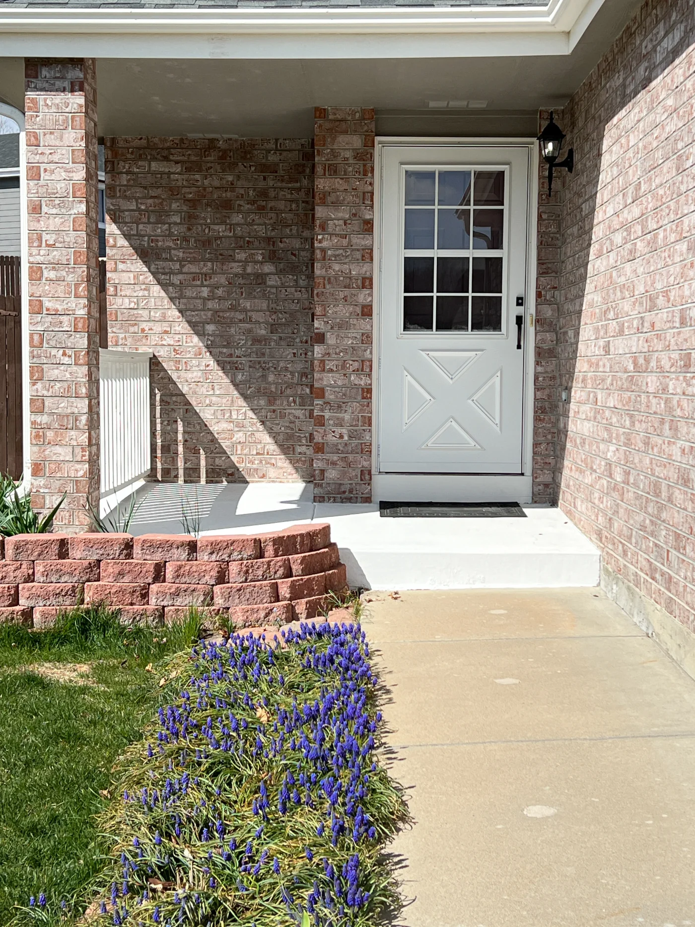 The front entryway of a red and tan brick home with multiple flower beds lining the walkway. The front door is white and has multiple square panes of glass. The part of the roof that can be seen is dark gray shingles.