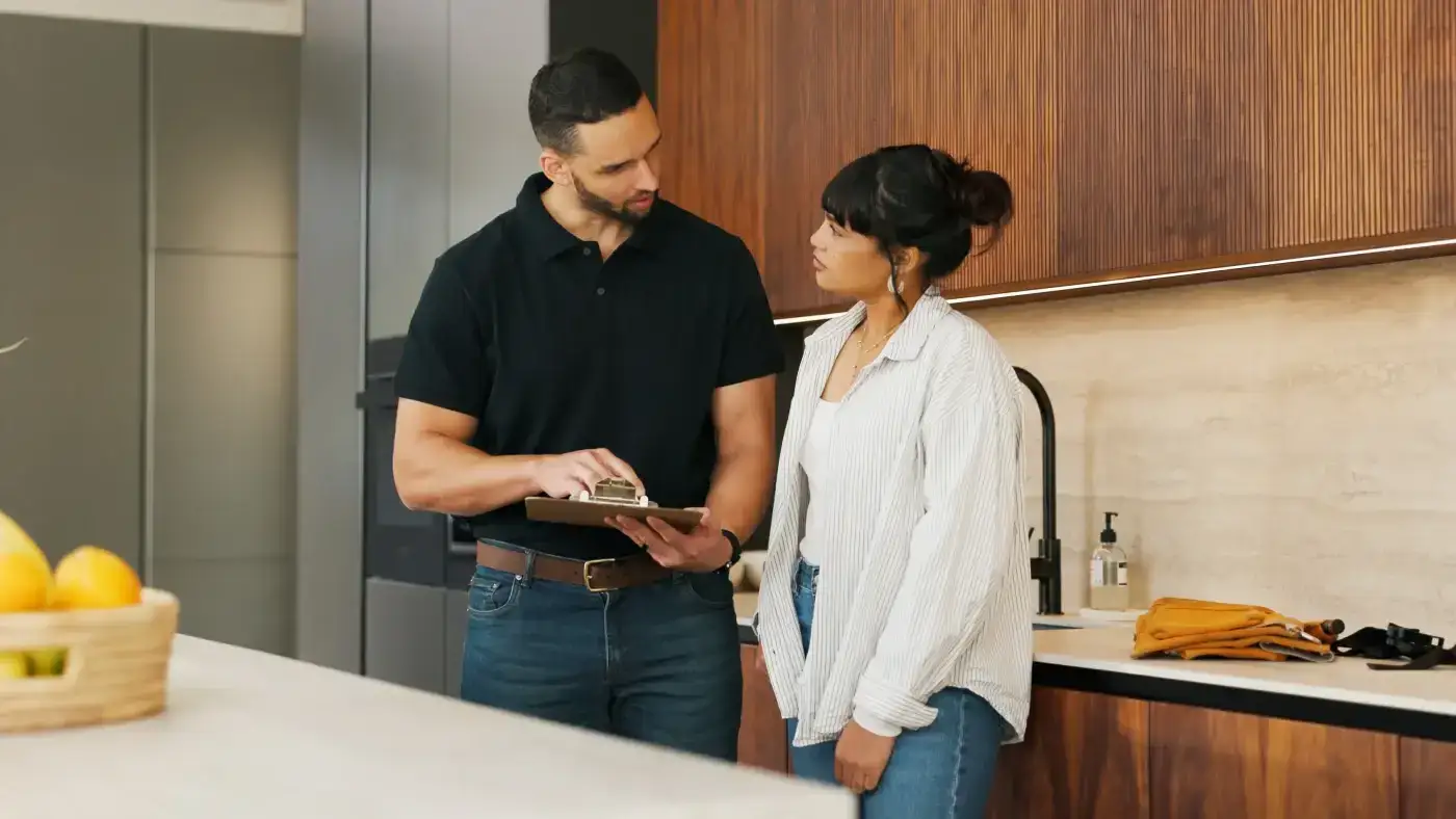 A man with a clipboard in hand holds a conversation with a woman in her kitchen.