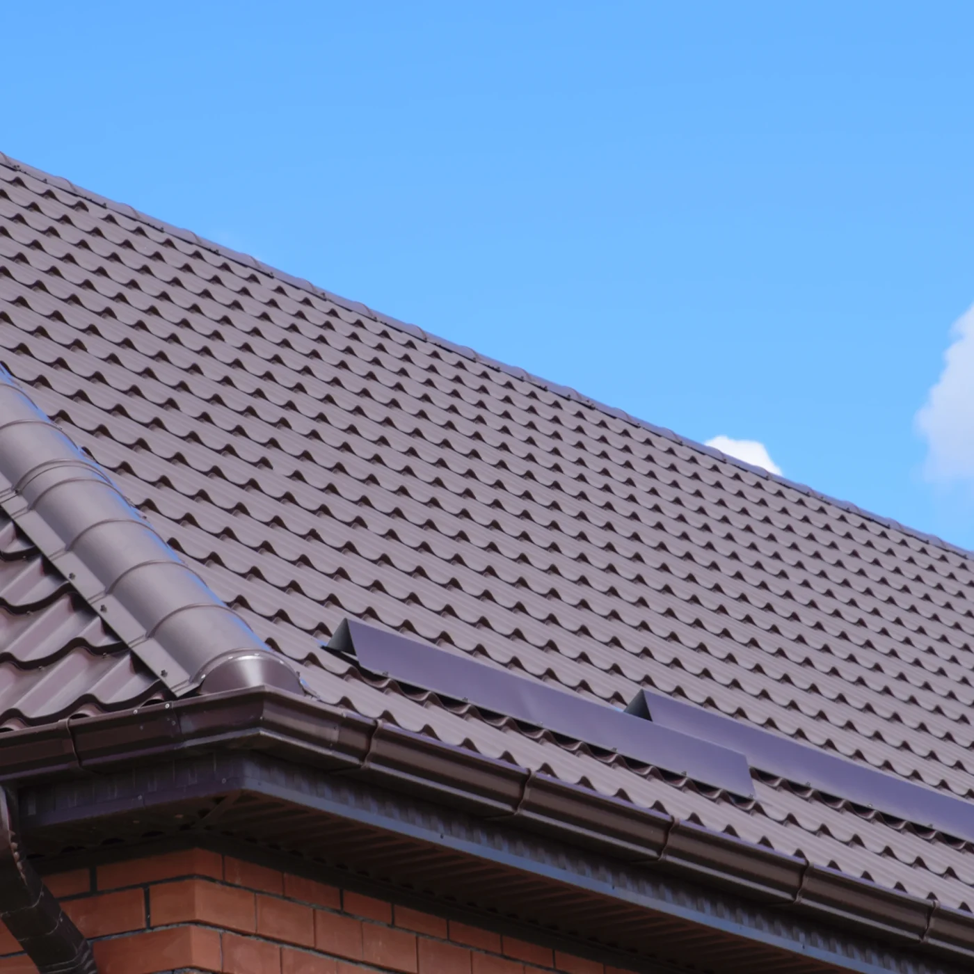 Close-up shot of a roof with dark brown metal shingles atop a red brick home. The sky above the roof is bright blue.